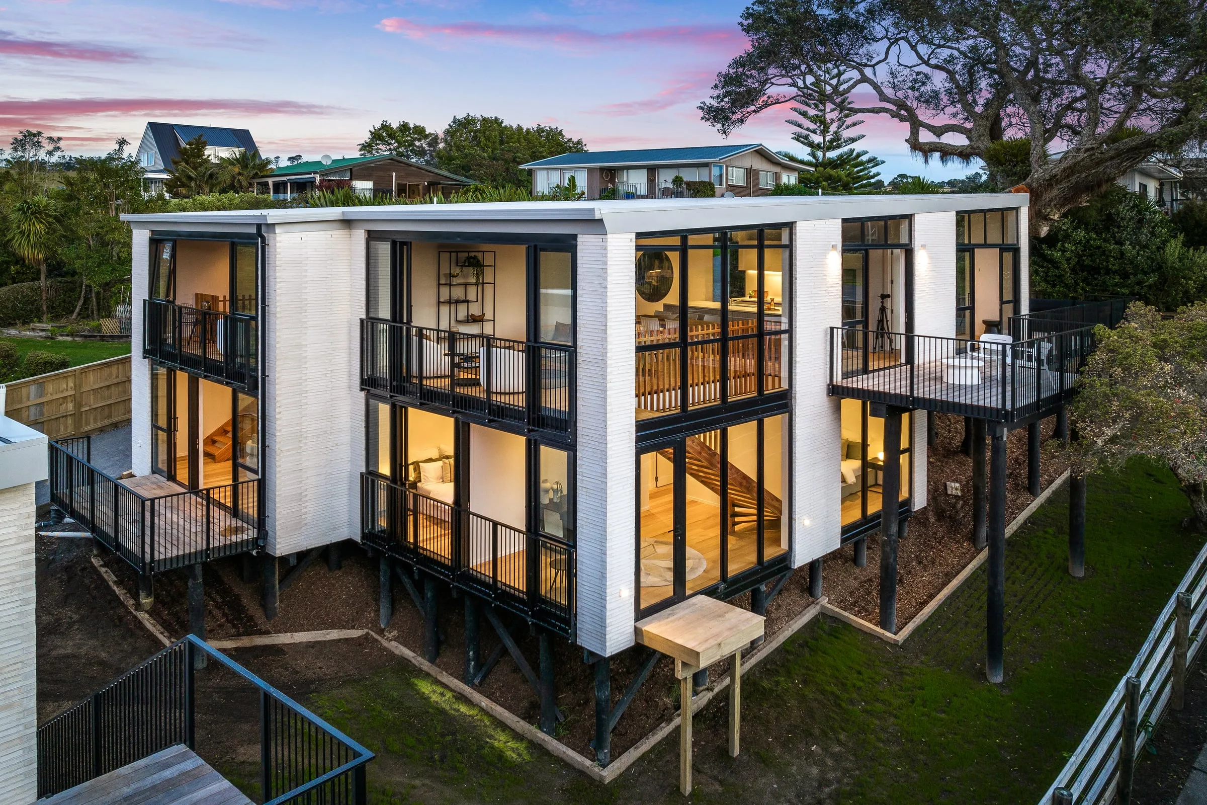A two-storey new build glazed across both levels with black aluminium windows and doors, lit from inside at dusk